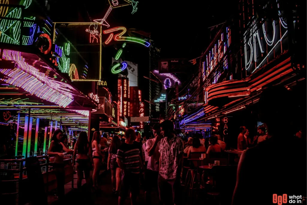 A vibrant street scene in Berlin at night, illuminated by colorful neon lights from bars and clubs. Crowds of people fill the bustling nightlife district, creating an energetic atmosphere with reflections of red, green, and purple lights dancing off glass and metal surfaces. The image captures the lively, eclectic spirit of Berlin's renowned party culture.
