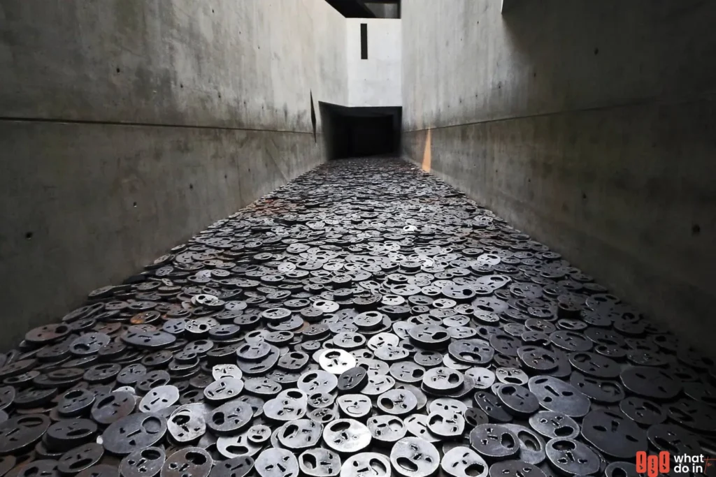 A haunting installation at the Jewish Museum in Berlin, featuring thousands of iron faces scattered across the floor of a narrow concrete corridor. The faces, each with hollow eyes and mouths, represent the victims of violence and oppression, creating a powerful atmosphere of remembrance and reflection.