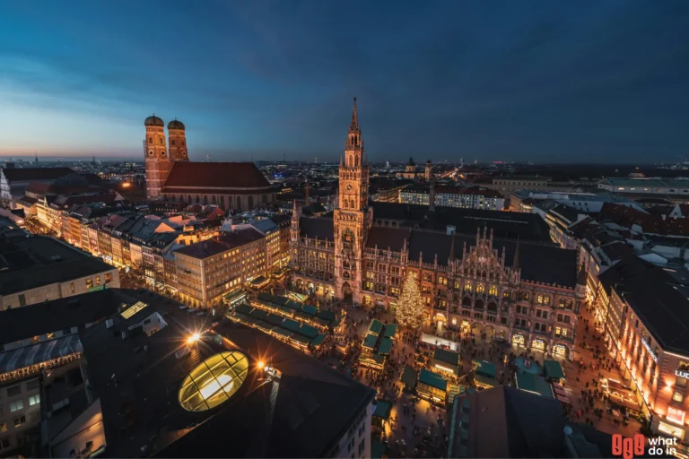 Aerial view of Munich's Marienplatz at dusk, beautifully illuminated with festive Christmas market stalls, a large decorated Christmas tree in front of the New Town Hall (Neues Rathaus), and the iconic twin towers of the Frauenkirche visible in the background.