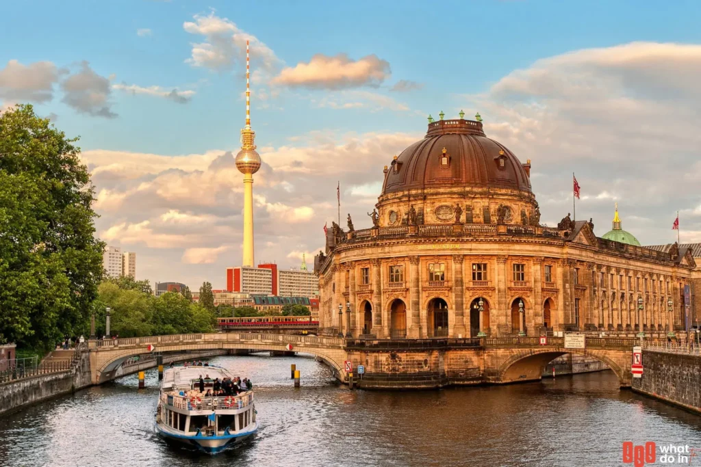A scenic view of Museum Island in Berlin at sunset, featuring the historic Bode Museum on the Spree River with a tour boat passing underneath. In the background, the iconic Berlin TV Tower (Fernsehturm) rises against a colorful sky with soft clouds. The scene captures the blend of cultural heritage, architectural beauty, and urban charm in the heart of Berlin.