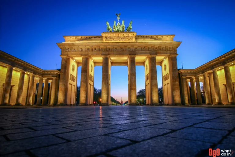 Illuminated Brandenburg Gate in Berlin at twilight, viewed from a low angle with a glowing blue and orange sky in the background. The neoclassical monument is highlighted with warm lighting, showcasing its iconic columns and the Quadriga statue on top.