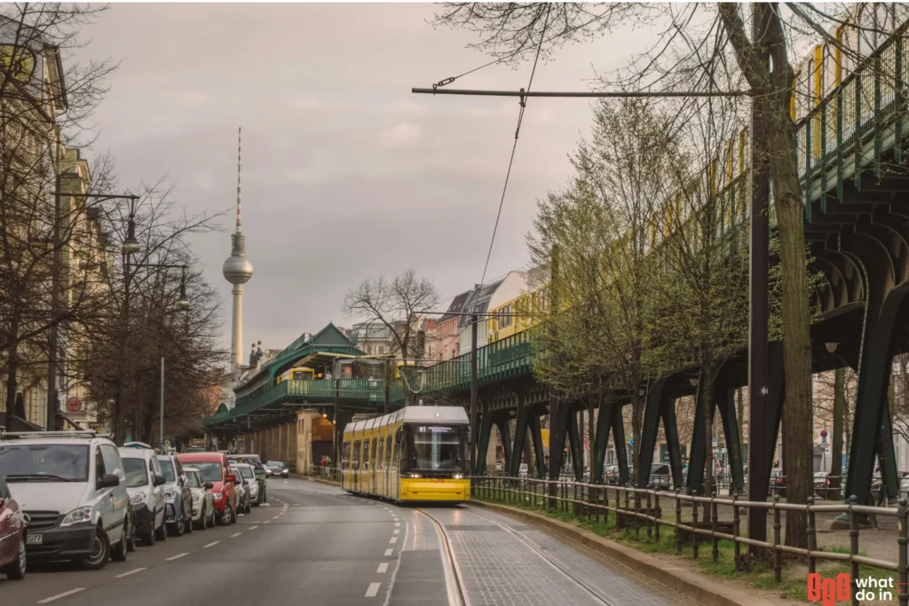 A street scene in Berlin featuring a yellow tram on a curved track and an elevated U-Bahn train passing above on green iron tracks. The iconic Fernsehturm (TV Tower) stands in the background, symbolizing the city's skyline. Lined with parked cars and early spring trees, the urban setting reflects Berlin's vibrant and efficient public transport system.