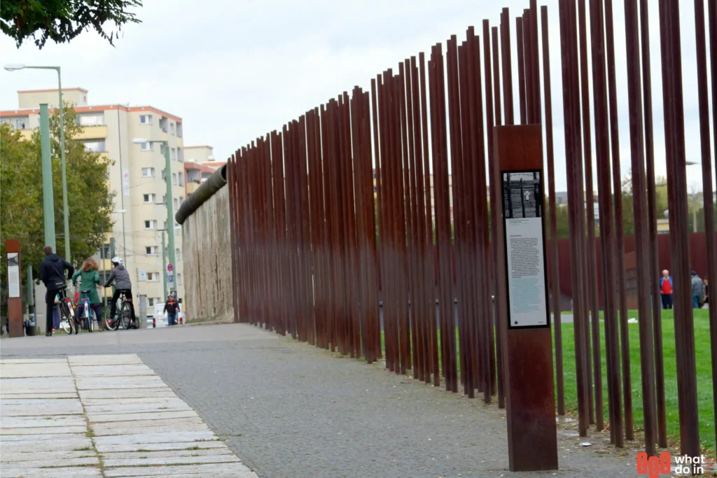 Visitors at the Berlin Wall Memorial, standing near preserved sections of the wall and rusted steel poles symbolizing the former border. A historical information panel is displayed in the foreground, with residential buildings and cyclists in the background.